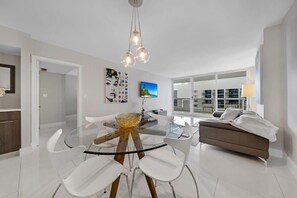 A modern living room with a glass dining table, white chairs, a brown sofa, and a large window offering a city view.