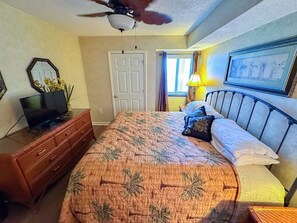 A bedroom with a bed, a dresser, a television, a ceiling fan, and a window with curtains.