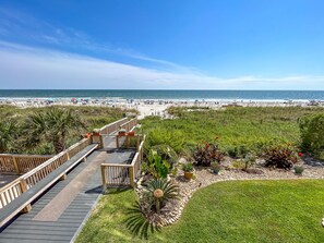 A beach view with a wooden boardwalk leading to the shore, surrounded by greenery and a clear sky.