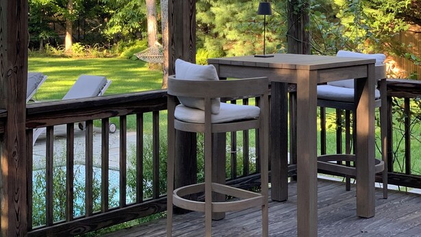 Een houten terras met een tafel en stoelen, omgeven door een houten hek, met uitzicht op een zwembad en weelderige begroeiing.