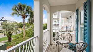 A wooden balcony with white railings, two wicker chairs with cushions, and a view of a white building with blue shutters. - Stay directly on 30A! Beautiful Beach House with private courtyard and panoramic views! Short walks to beach, pool, Rosemary and Alys! (Panama City Beach)