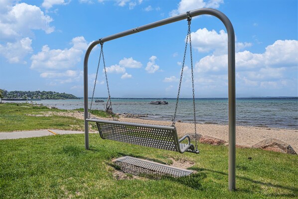 Un banco de parque con estructura metálica y cadenas, situado cerca de una playa con vistas al agua y tierra a lo lejos.