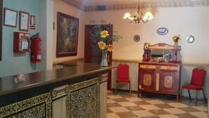 Un área de recepción con suelo a cuadros, un mostrador de recepción, dos sillas rojas, un jarrón con flores, un extintor, certificados enmarcados y un cuadro en la pared.