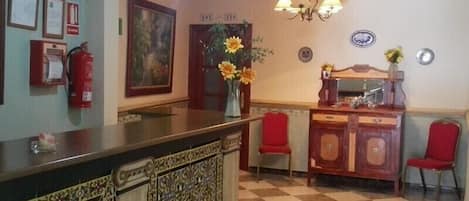 Un área de recepción con suelo a cuadros, un mostrador de recepción, dos sillas rojas, un jarrón con flores, un extintor, certificados enmarcados y un cuadro en la pared.