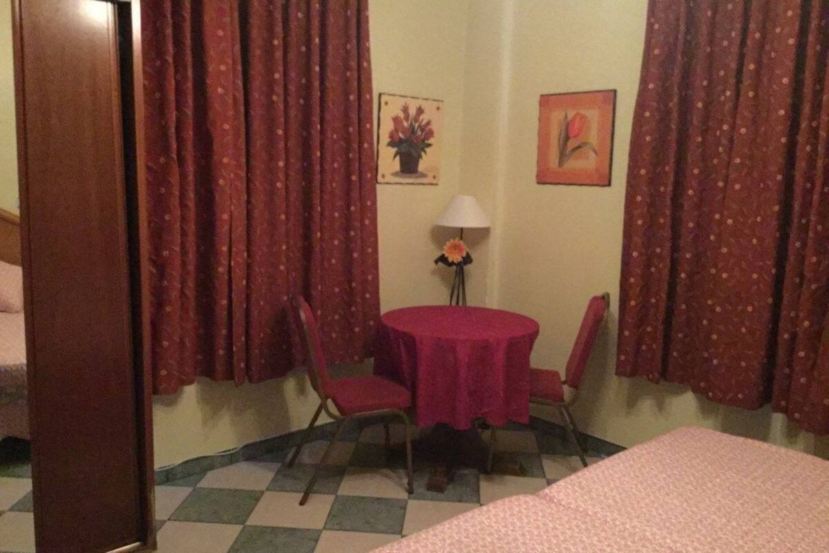 Una habitación con una cama, una mesa con sillas, una lámpara y dos cuadros en la pared.