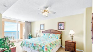 A bedroom with a bed, a nightstand, a ceiling fan, a window with curtains, and a view of the ocean.