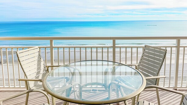A balcony with a glass table and chairs overlooking a beach.