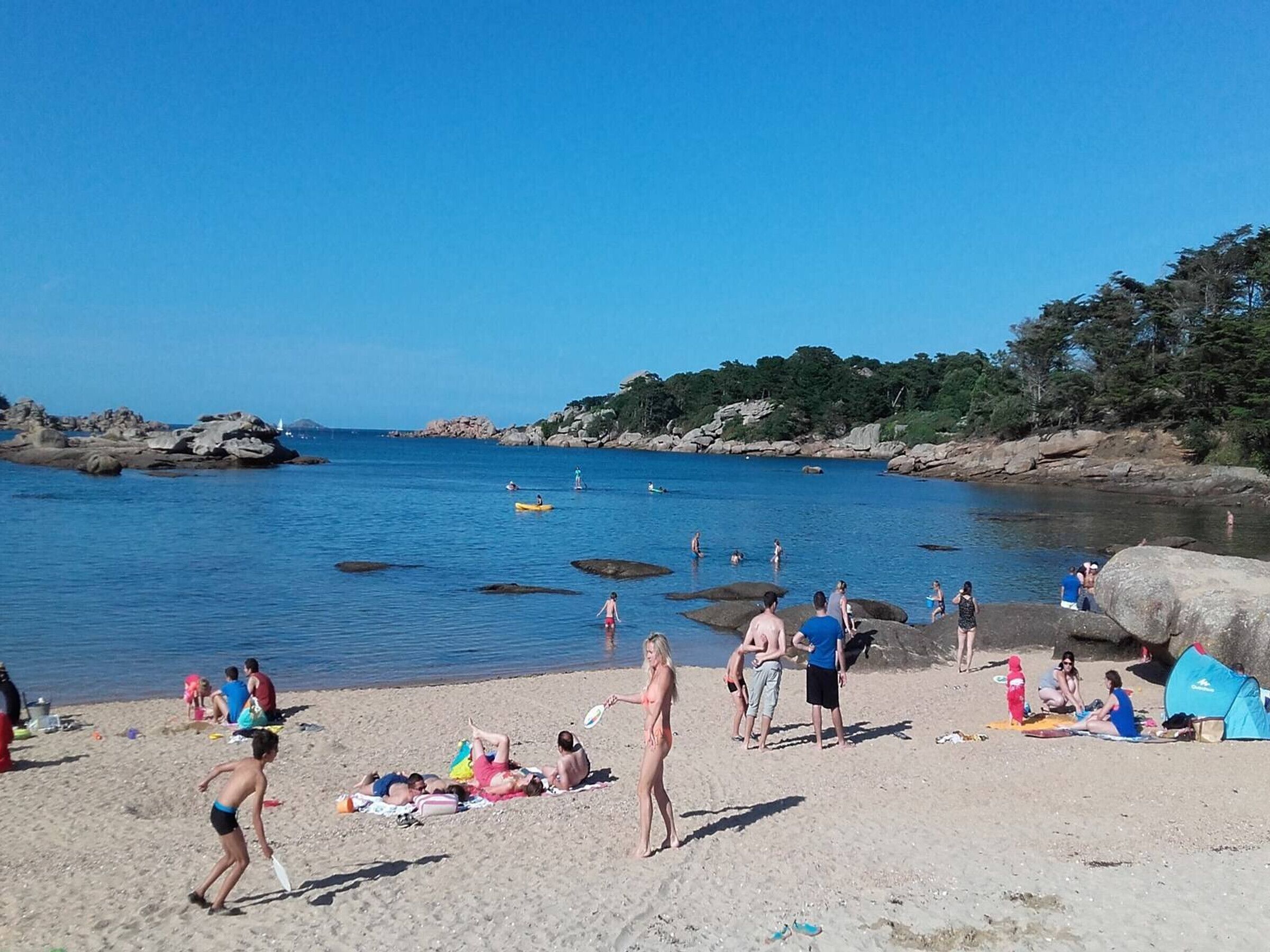 A beach with people sunbathing and swimming, rocky outcrops, and a clear blue sky.