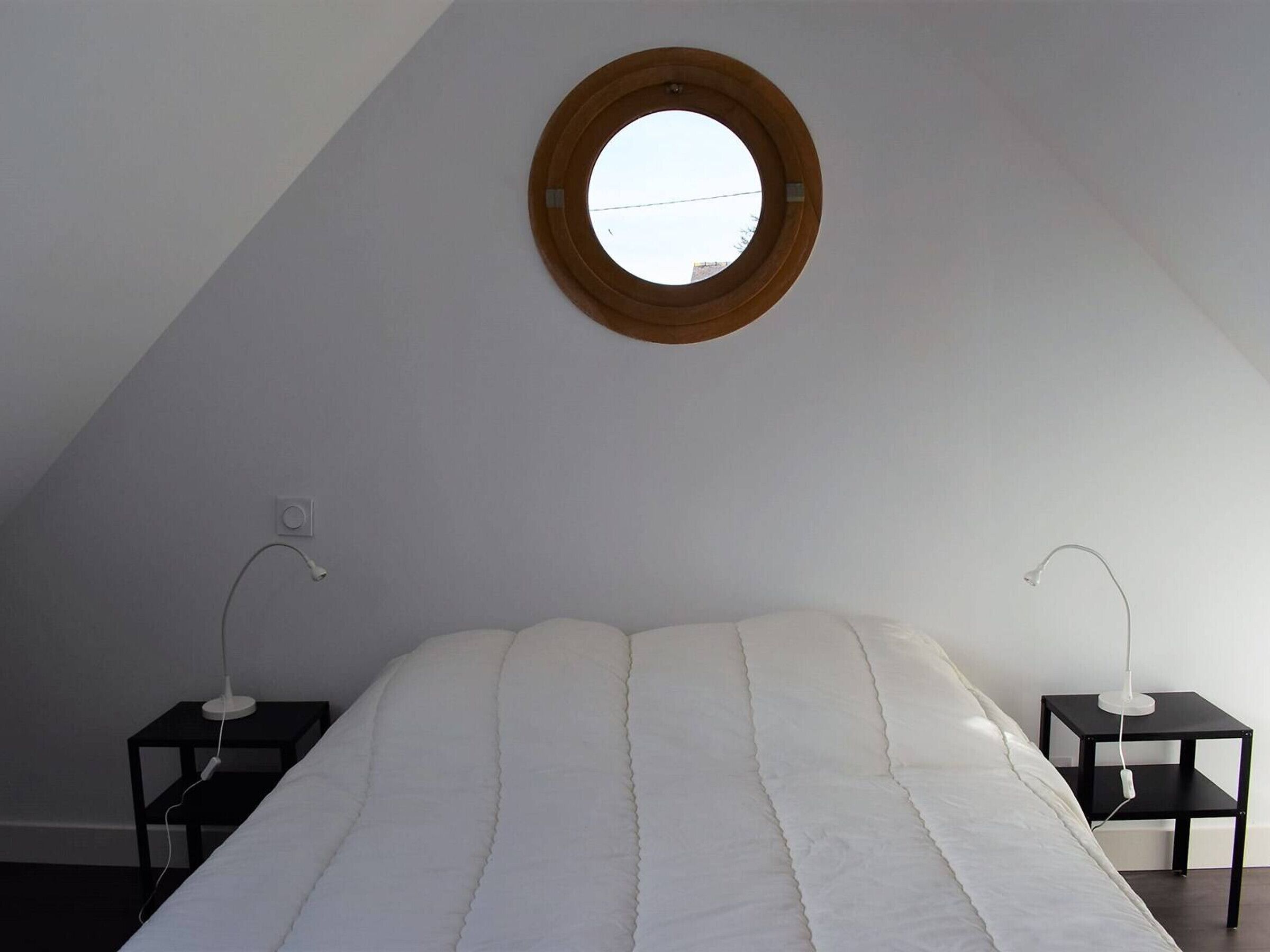 A bedroom with a sloped ceiling, a round window, a bed with white bedding, two bedside tables with lamps, and a small shelf on one table.