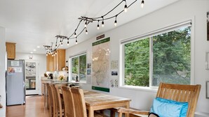 Una cocina moderna con armarios de madera, un comedor con mesa y sillas de madera, un refrigerador y un ventanal con vistas a árboles.