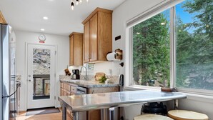 Una cocina moderna con armarios de madera, un refrigerador de acero inoxidable, un horno empotrado y un amplio ventanal con vista a árboles.