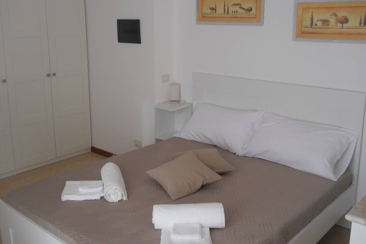 A neatly made bed with white and beige pillows, a small bedside table with a lamp, and a framed picture on the wall.
