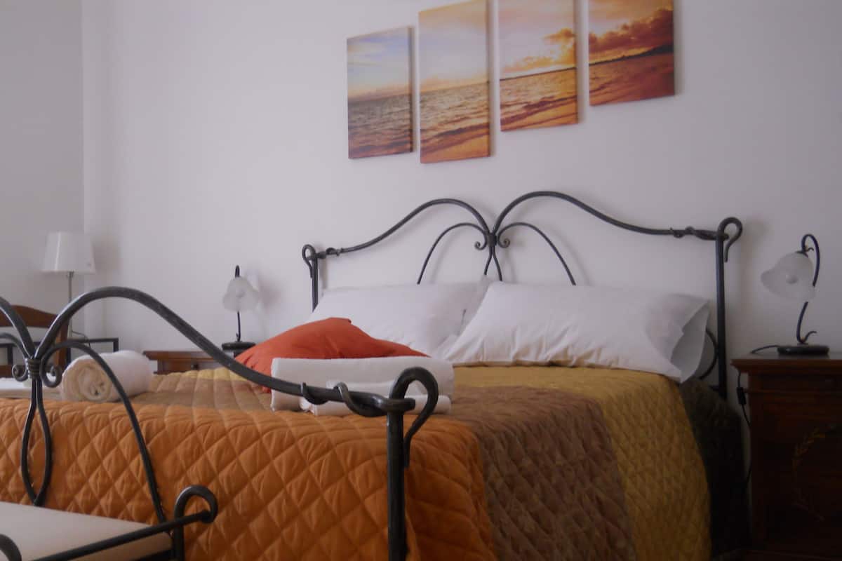 A neatly made bed with a quilted orange and brown cover, a wooden nightstand with a lamp, and three sunset paintings on the wall.