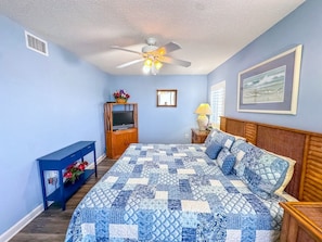 Quarto com cama, ventilador de teto, televisão, mesa de cabeceira e um quadro na parede.
