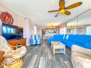 A spacious living room with a large blue sofa, a wooden coffee table, and a television. There is a ceiling fan and a decorative wall hanging. - North Shore Villas 801! Coastal Dream Stay – 4BR Oceanfront NMB Escape! (North Myrtle Beach)