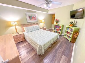 A bedroom with a bed, bedside tables, lamps, a bookshelf, and a television. - North Shore Villas 801 (North Myrtle Beach)