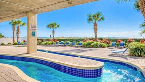 Zona de piscina con bordes de azulejos azules, sillas de descanso, palmeras y un cartel que prohíbe el clavado.