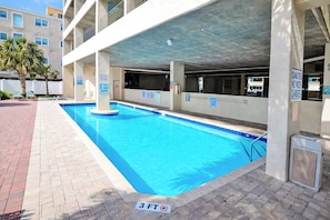 A swimming pool with a 3 FT depth limit sign, surrounded by a brick walkway, under a covered parking area with multiple levels. - North Shore Villas 801 (North Myrtle Beach)