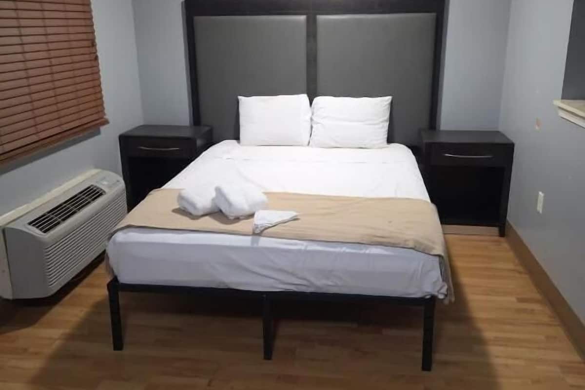 A hotel room with a bed, two bedside tables, a heater, and a window with blinds.