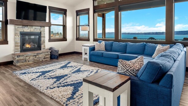 Ein Wohnzimmer mit Kamin aus Stein, einem blauen Sofa und großen Fenstern mit Blick auf einen See.
