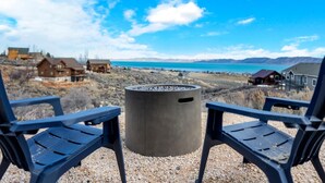 Blick auf einen See mit Häusern im Hintergrund, einer Beton-Feuerstelle und zwei Stühlen, die zur Feuerstelle zeigen.