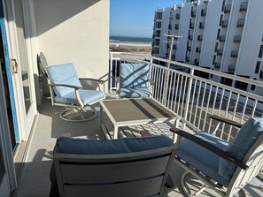 A balcony with a view of the beach, white railing, and two blue chairs.
