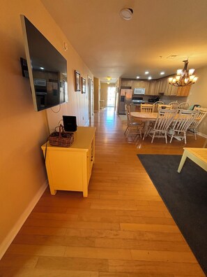 A modern kitchen with wooden cabinets, a dining area with a table and chairs, and a hallway with a flat-screen TV mounted on the wall.