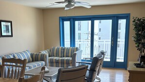 Uma área de refeições com mesa e cadeiras de madeira, ventilador de teto e vista através de uma janela com moldura azul.