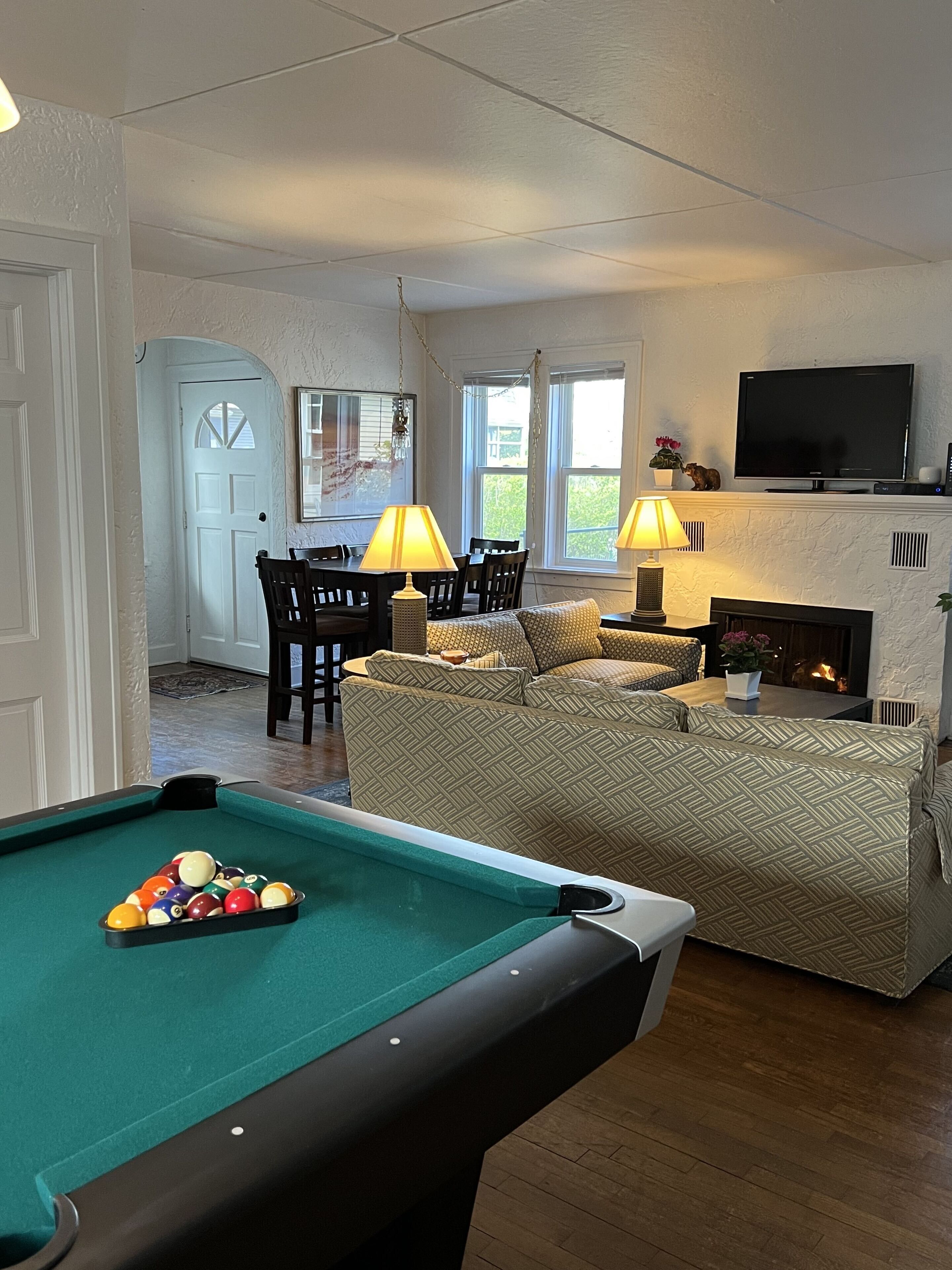 Une table de billard avec des billes, un canapé, un téléviseur et une cheminée dans une pièce au plancher en bois et aux murs blancs.