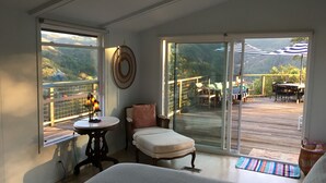 A bedroom with a large window, a bed, a chair, a table, and a view of a deck with outdoor furniture.