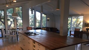 A spacious kitchen with a large island, white cabinetry, and a view of trees through large windows.