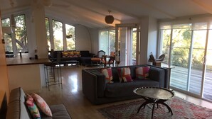 A spacious living area with a sofa, a coffee table, and a rug. There is a kitchen area with a bar counter and a dining table with chairs. Large windows provide natural light and views of the outdoors.