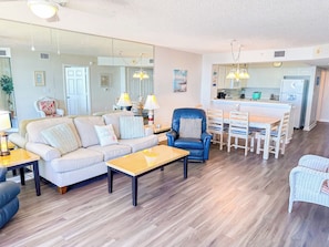 A modern living room with a sofa, coffee table, and armchairs, a dining area with a wooden table and chairs, and a kitchen with white cabinets and a refrigerator.