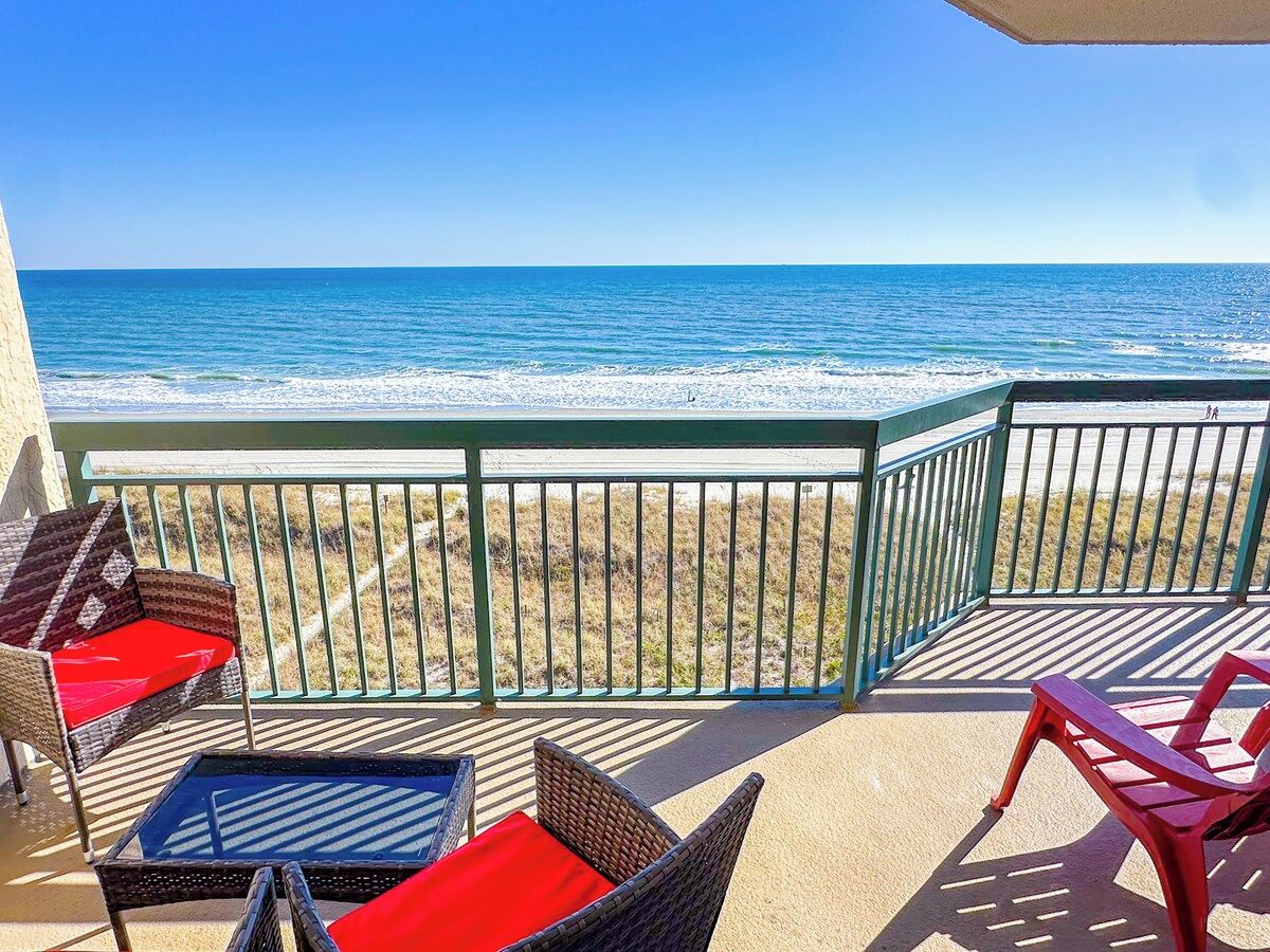 A balcony with wicker chairs and a table, overlooking a beach with waves.