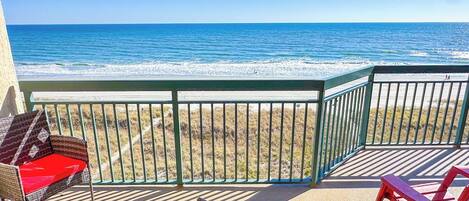 A balcony with wicker chairs and a table, overlooking a beach with waves.