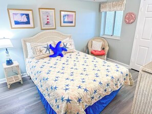 Quarto com uma cama grande, uma mesa de cabeceira, uma cadeira de vime e uma janela com cortinas.
