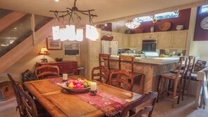 Ein Essbereich mit Holztisch, Stühlen und einer Obstschale. Im Hintergrund ist eine Küche mit Schränken und einem Kühlschrank zu sehen.