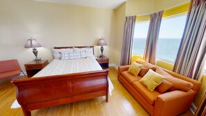 A bedroom with a wooden bed, a sofa, two bedside tables with lamps, and a view of the sea through curtains.