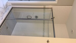 Une douche fermée par des parois de verre, avec une structure métallique, des murs recouverts de carreaux blancs et une fenêtre située en hauteur.