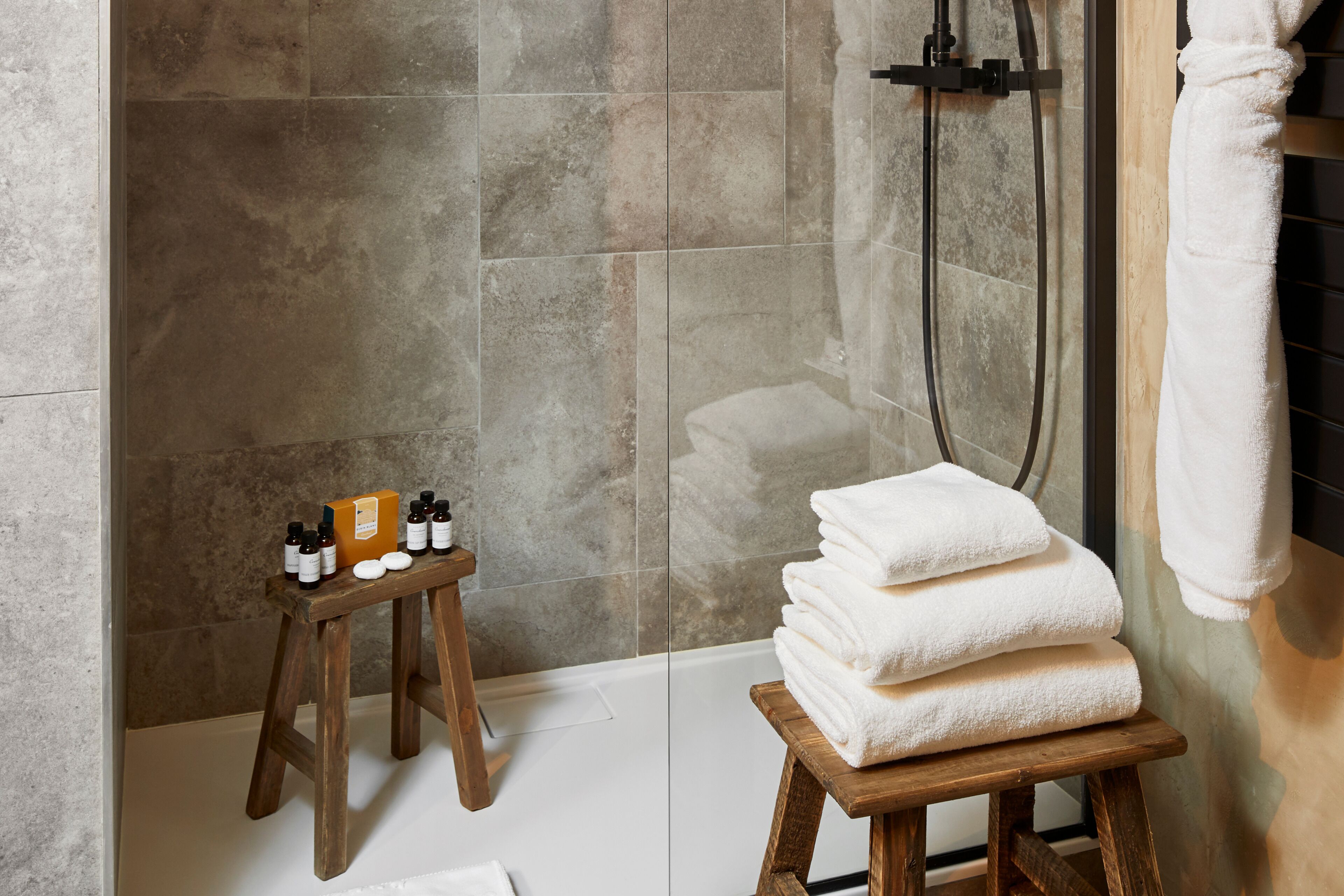 A bathroom with a shower, a wooden stool with toiletries, and a stack of towels.