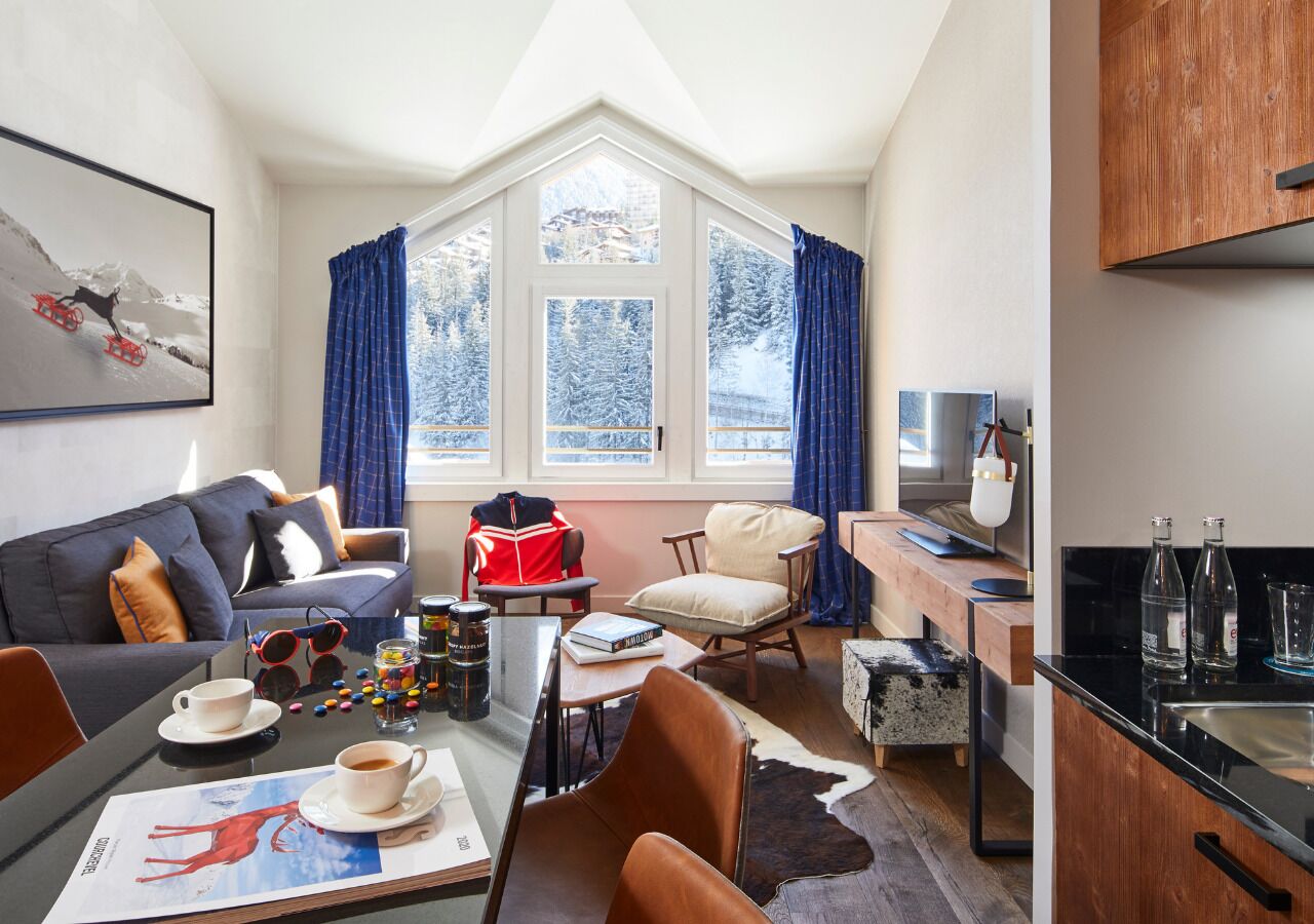 A cozy living room with a sofa, coffee table, and a dining area with a table and chairs. A large window with curtains is visible, and a kitchen area with a sink and cabinets is also present.