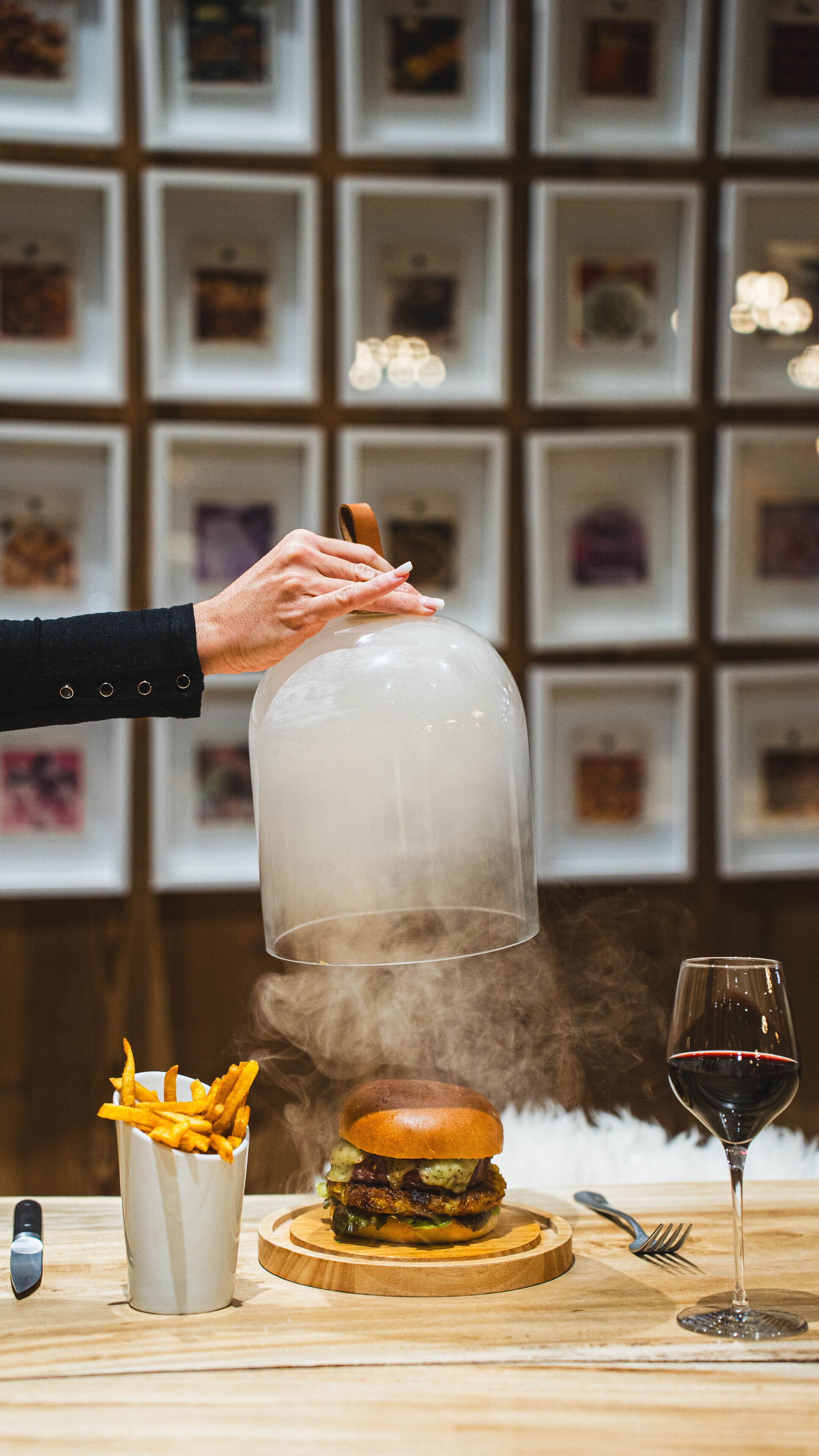 A person holding a transparent dome over a burger, with fries and a wine glass on the table.