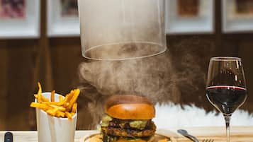 Une personne tenant un dôme transparent au-dessus d’un hamburger, avec des frites et un verre de vin sur la table.