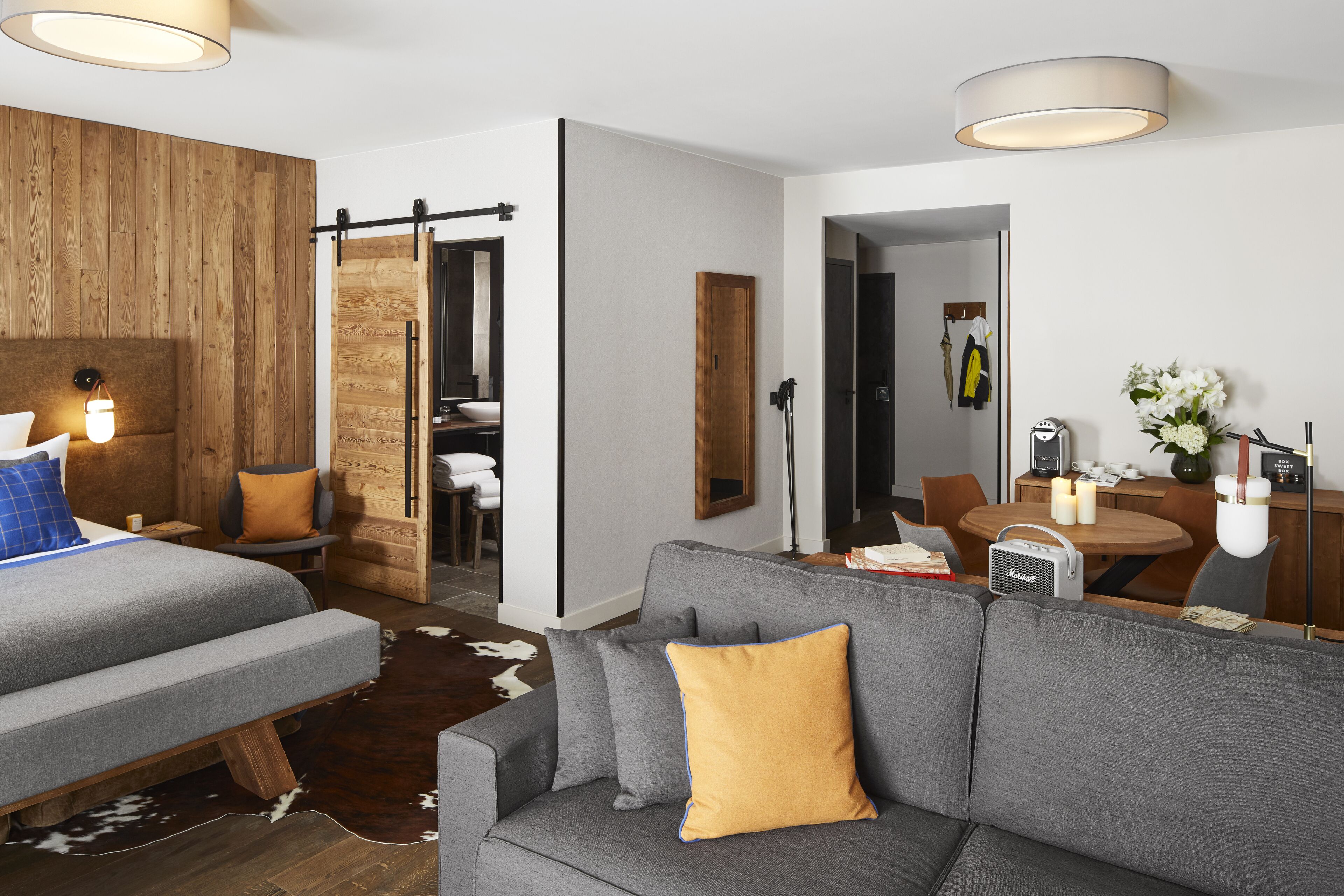 Una habitación de hotel moderna con un sofá gris, detalles de madera, un tapiz de piel de vaca, una zona de comedor con mesa y sillas, y un dormitorio independiente con cama.