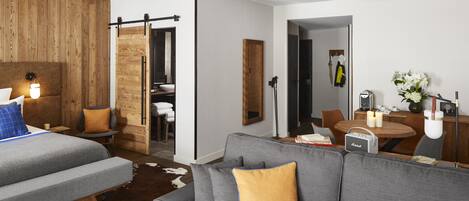 Una habitación de hotel moderna con un sofá gris, detalles de madera, un tapiz de piel de vaca, una zona de comedor con mesa y sillas, y un dormitorio independiente con cama.
