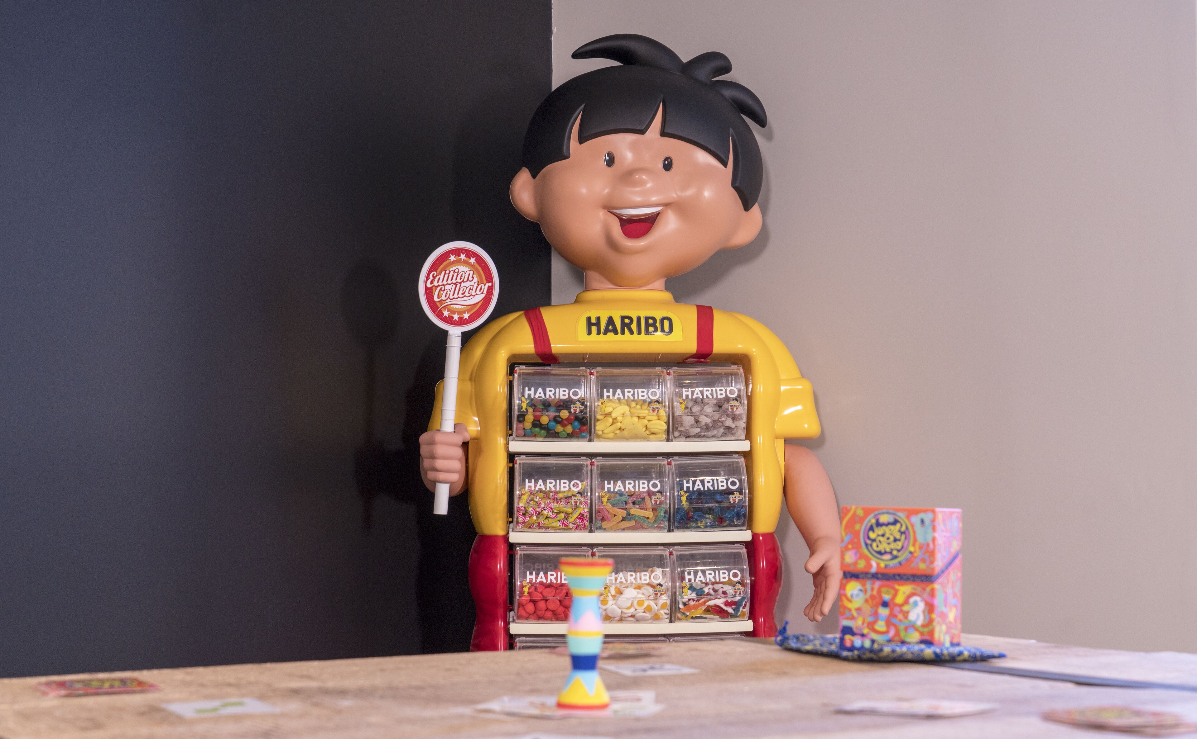 Una figura de juguete de un personaje sosteniendo una piruleta y de pie detrás de una exhibición de caramelos Haribo.