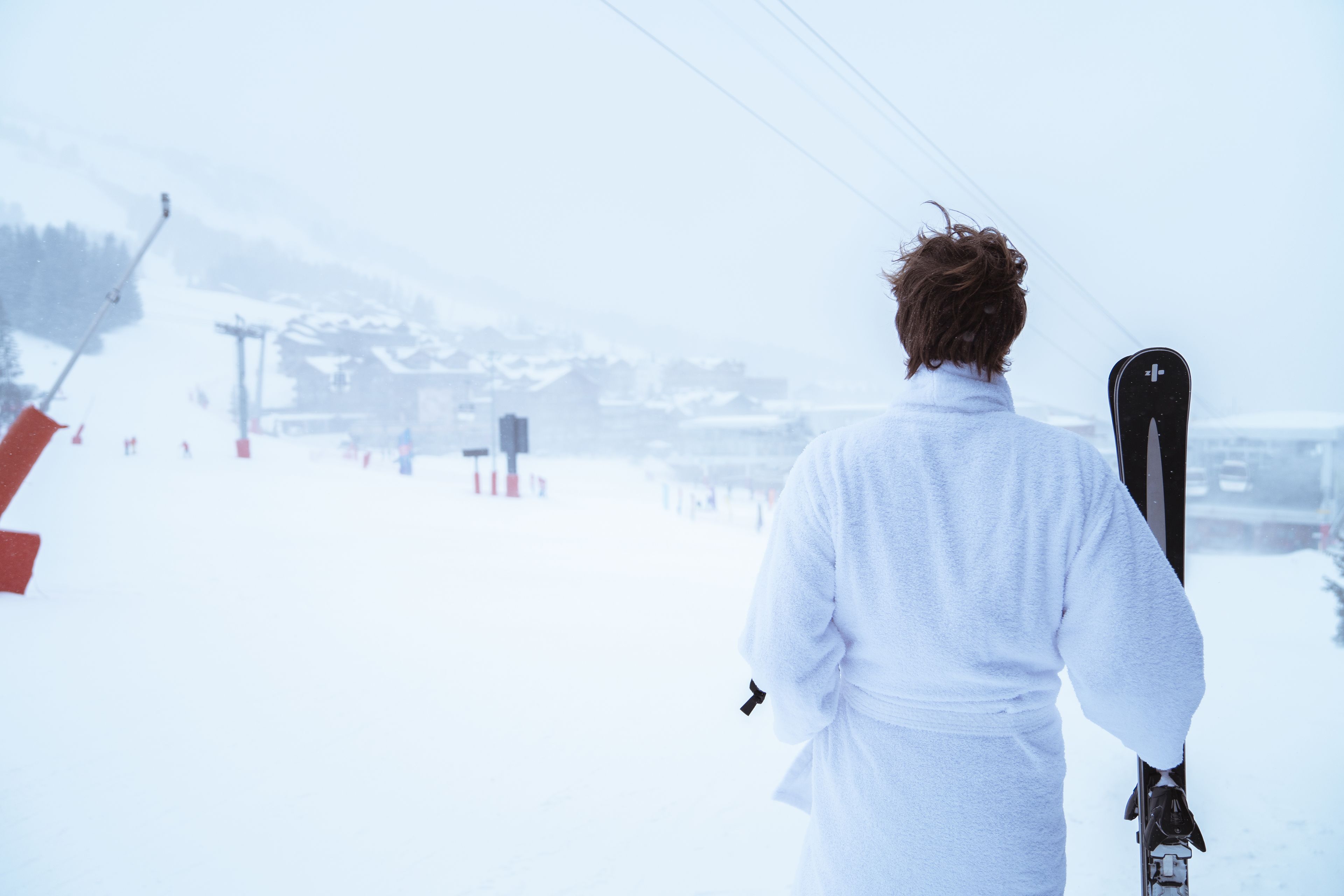 A person in a white robe holding skis, standing on a snowy slope with ski lifts in the background.