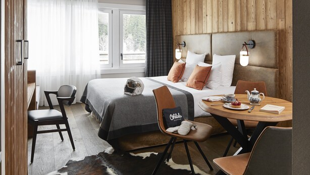 A modern hotel room with a large bed, a wooden headboard, a dining table with chairs, and a view of the outdoors through the windows. - Ecrin Blanc Resort Courchevel (Courchevel)