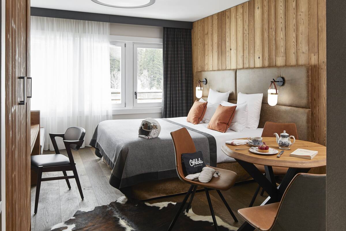 Una camera d'albergo moderna con un letto grande, una testiera in legno, un tavolo da pranzo con sedie e vista all'esterno attraverso le finestre.