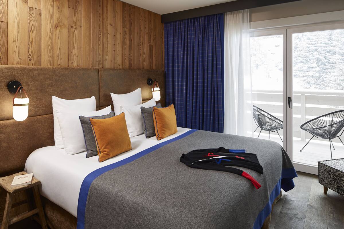 Camera d'albergo con testiera in legno, un letto grande con copripiumino grigio e blu, cuscini bianchi e marroni, un comodino con una lampada e vista su un balcone esterno innevato.